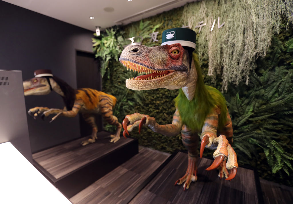 Dinossauros-robôs com quepe verde recepcionam hóspedes em hotel japonês.