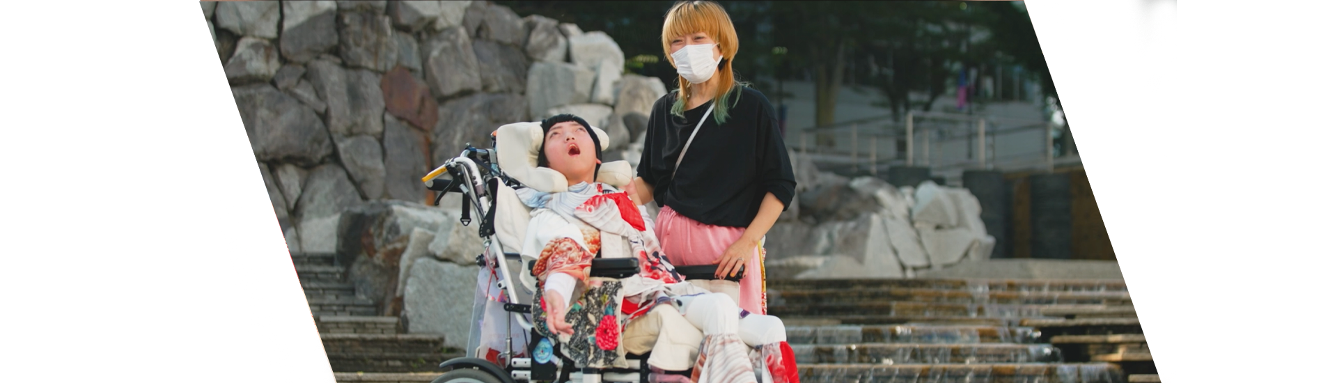 Na imagem, está uma jovem menina japonesa, em uma cadeira de rodas especializada, sendo acompanhada pela sua mãe.O ambiente ao redor parece ser ao ar livre, com uma escadaria de pedras.