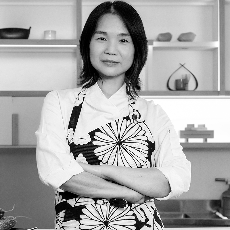 Foto em preto e branco de Telma Shiraishi, uma mulher com expressão confiante, olhando diretamente para a câmera. Ela veste um uniforme de chef e um avental com estampa floral, de braços cruzados, em uma cozinha com prateleiras ao fundo.