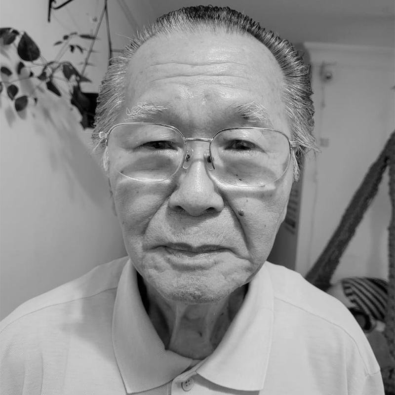 A imagem mostra Jorge Kinoshita, um homem idoso de feições asiáticas, usando óculos e vestindo uma camisa polo. Ele tem cabelos curtos, grisalhos, penteados para trás. A foto é em preto e branco, e ele olha diretamente para a câmera com uma expressão serena. Ao fundo, há uma parede clara com algumas plantas penduradas.