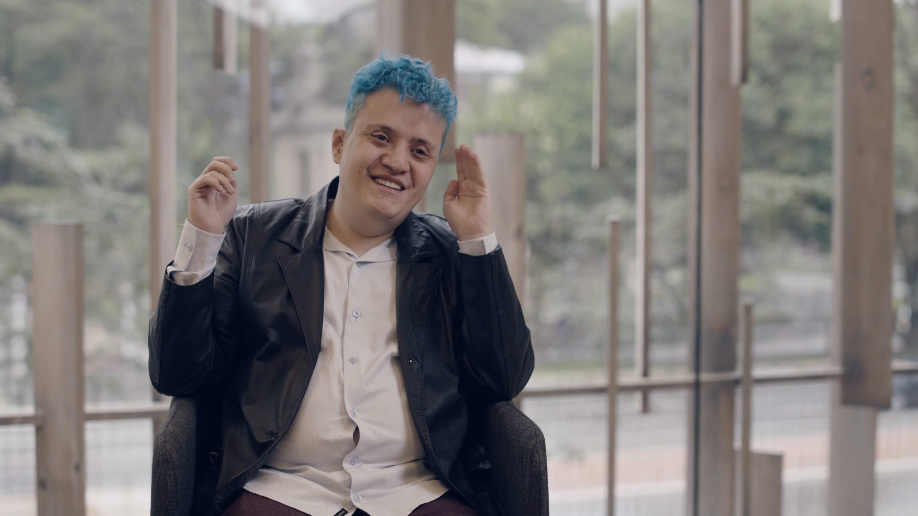 Foto de menino jovem, de cabelo azul, sentado no interior da JHSP. Ele sorri 